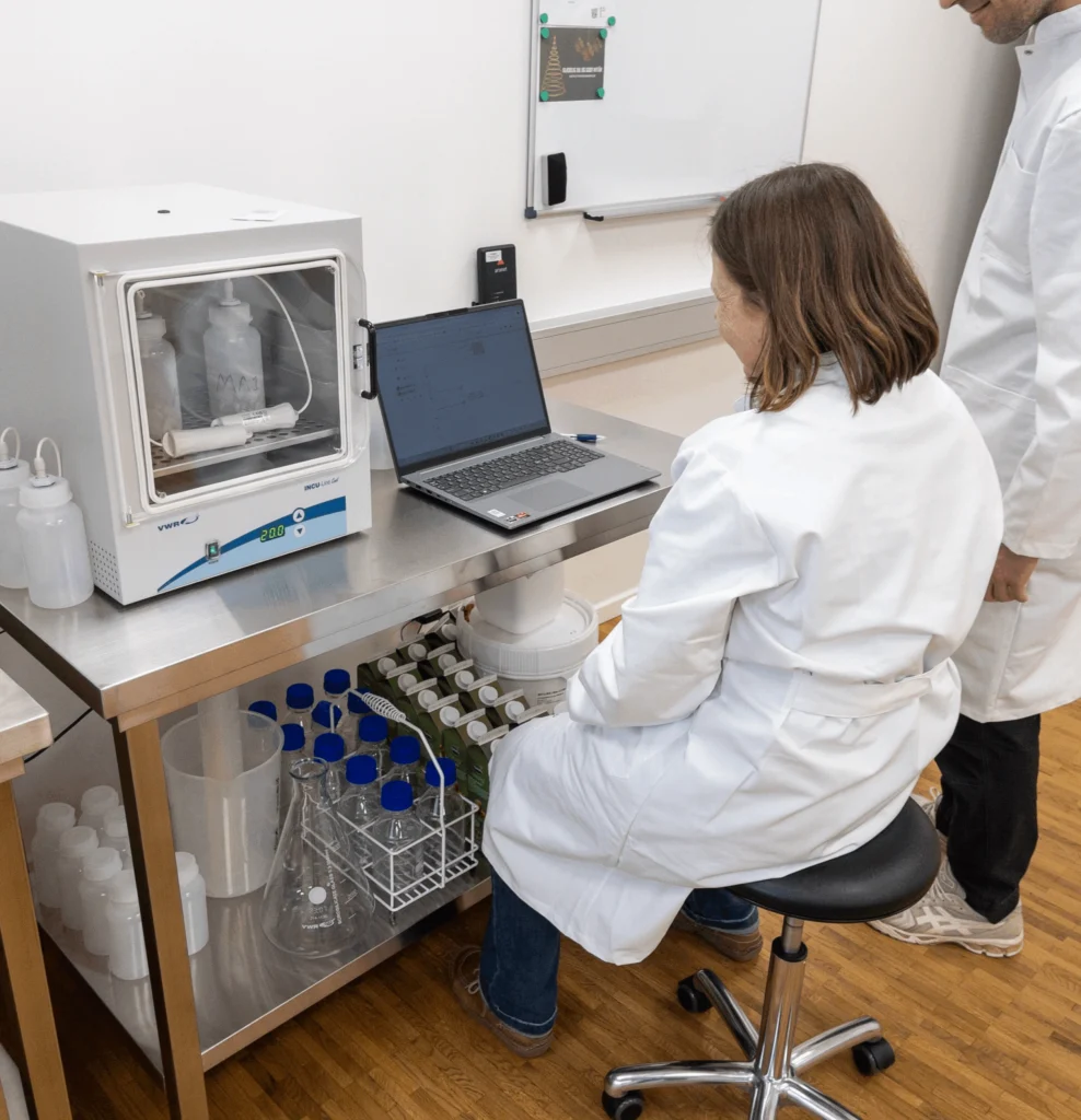 Laboratoriemedarbejdere arbejder med tørreskab og computer i moderne laboratoriemiljø med udstyr og flasker.
