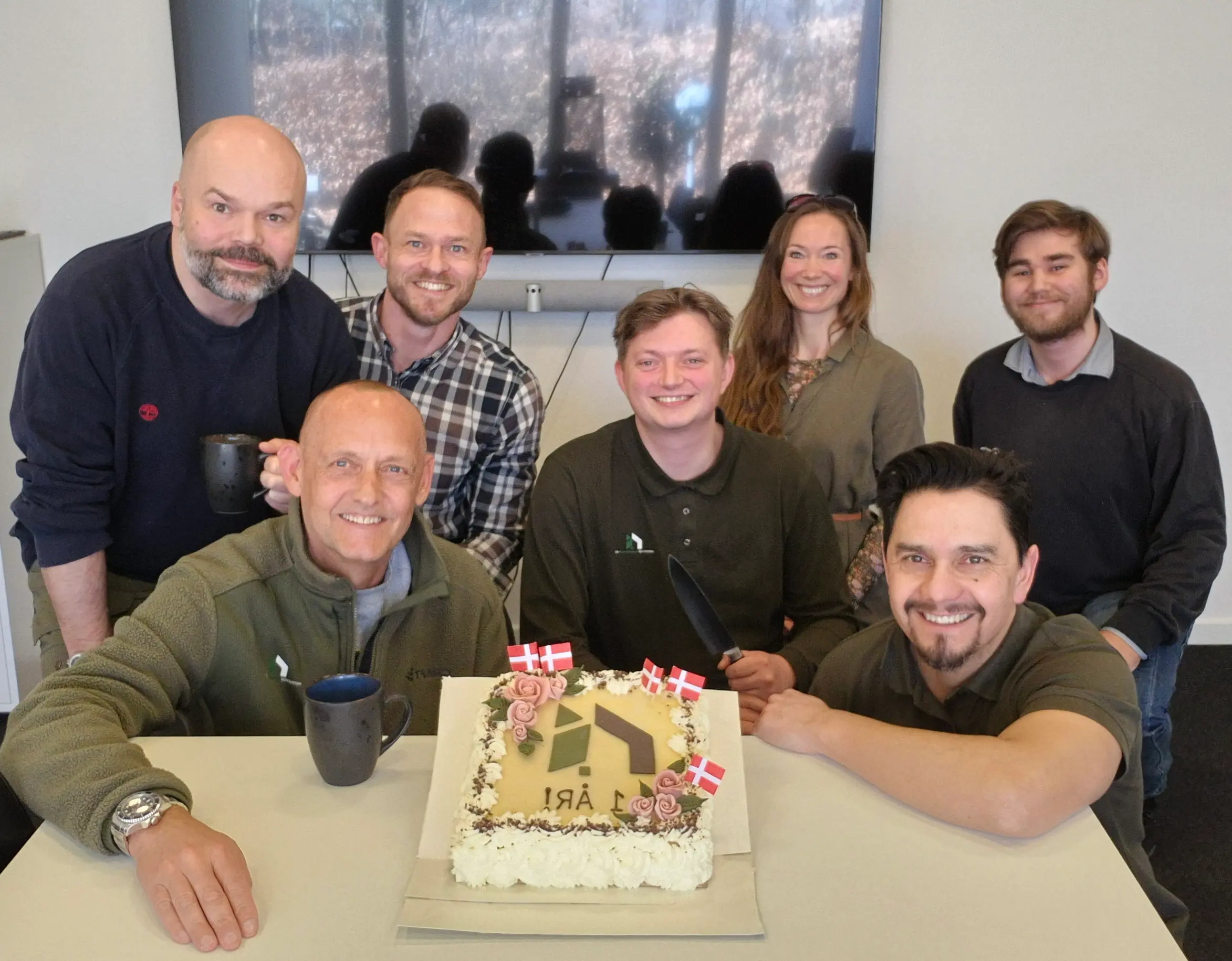 Gruppe af medarbejdere fra Institut for Bygningsmiljø fejrer virksomhedens 1-års jubilæum med kage dekoreret med logo og dannebrogsflag, smilende foran et mødebord.