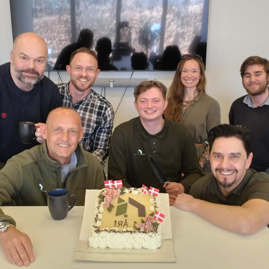 Gruppe af medarbejdere fra Institut for Bygningsmiljø fejrer virksomhedens 1-års jubilæum med kage dekoreret med logo og dannebrogsflag, smilende foran et mødebord.